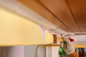A hand uses a paintbrush to apply yellow paint to exterior wood trim.